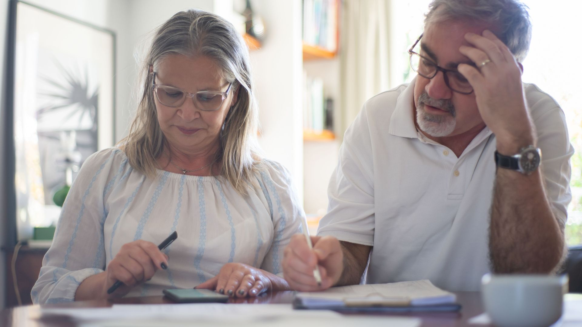 Senior couple calculating expenses at home office desk with documents and notes.