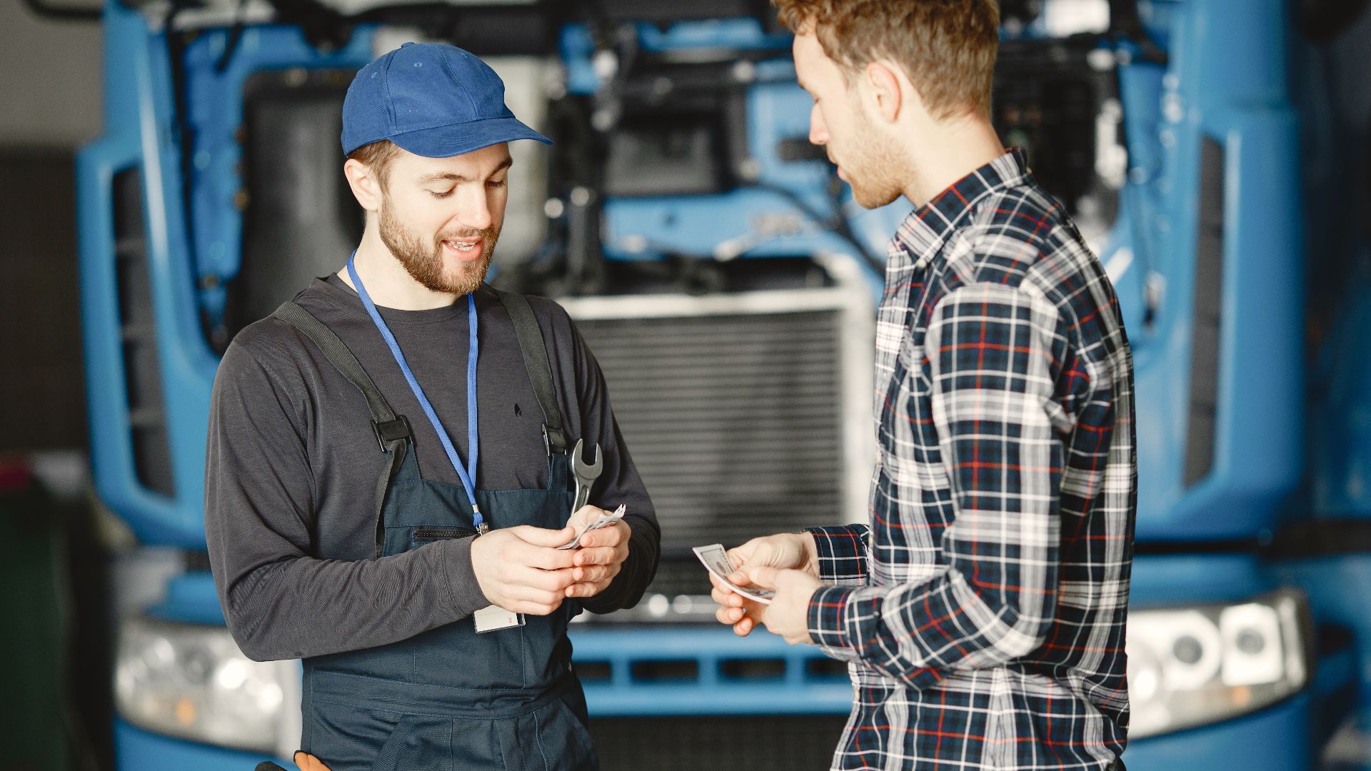 Auto mechanic in blue uniform receives payment from customer in a garage setting with a truck in the background.