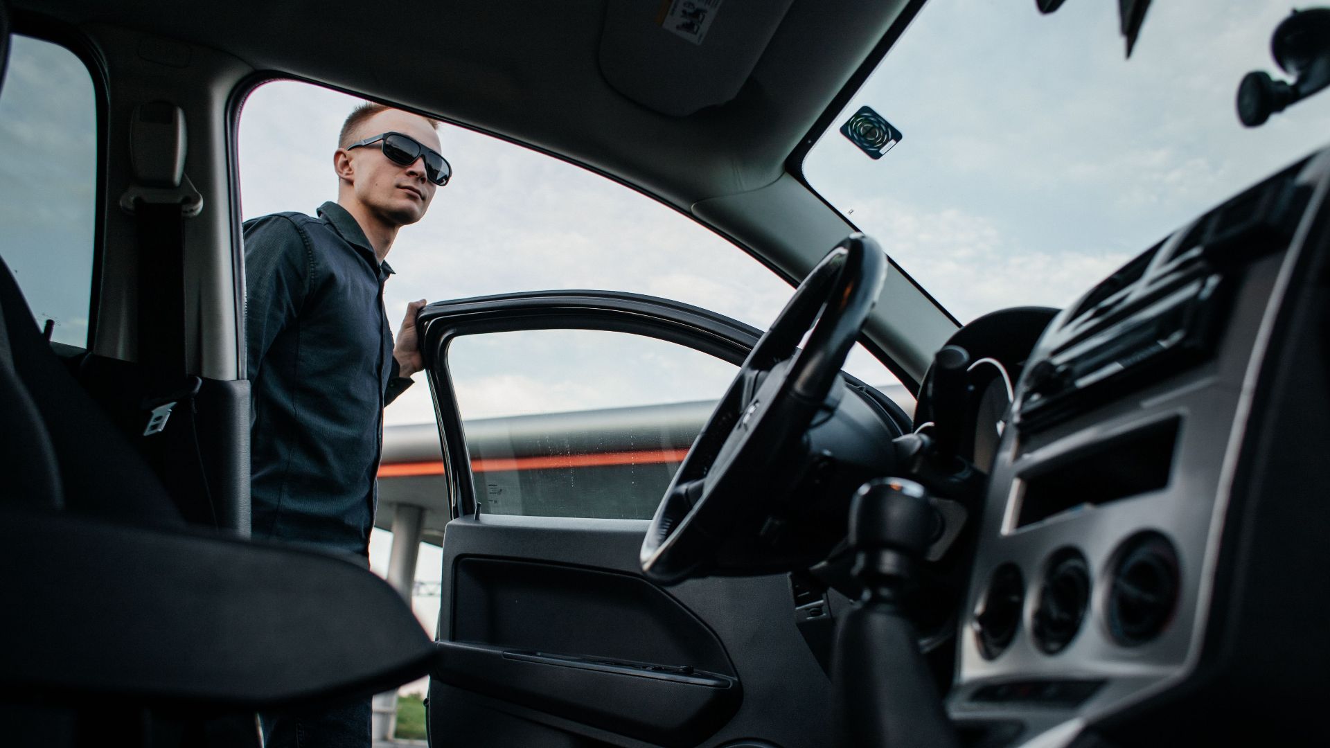A man in sunglasses stands by an open car door, viewed from the interior.