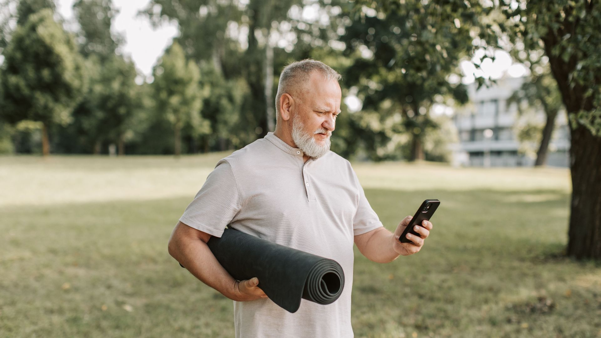 Elderly man with yoga mat texting on phone in a park.