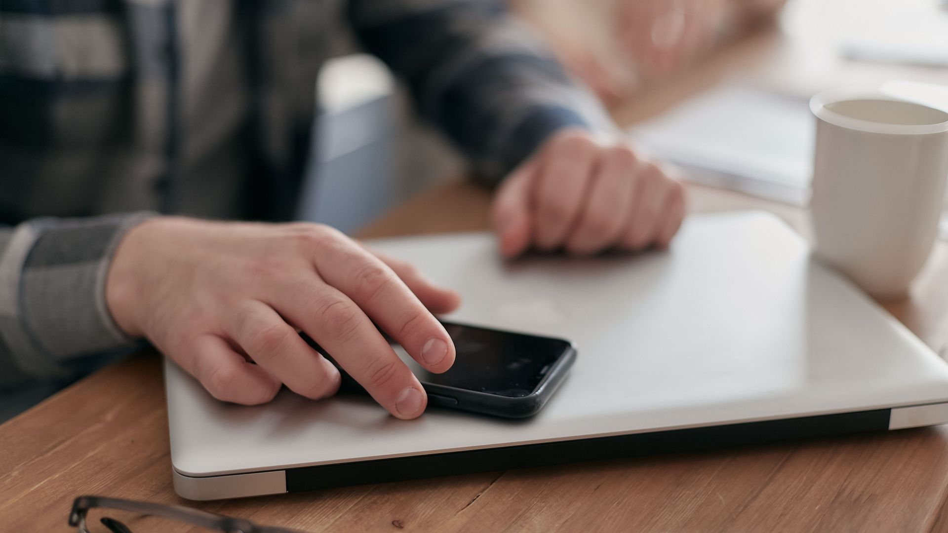 Close-up of hands using smartphone and laptop at a wooden desk with glasses and coffee.
