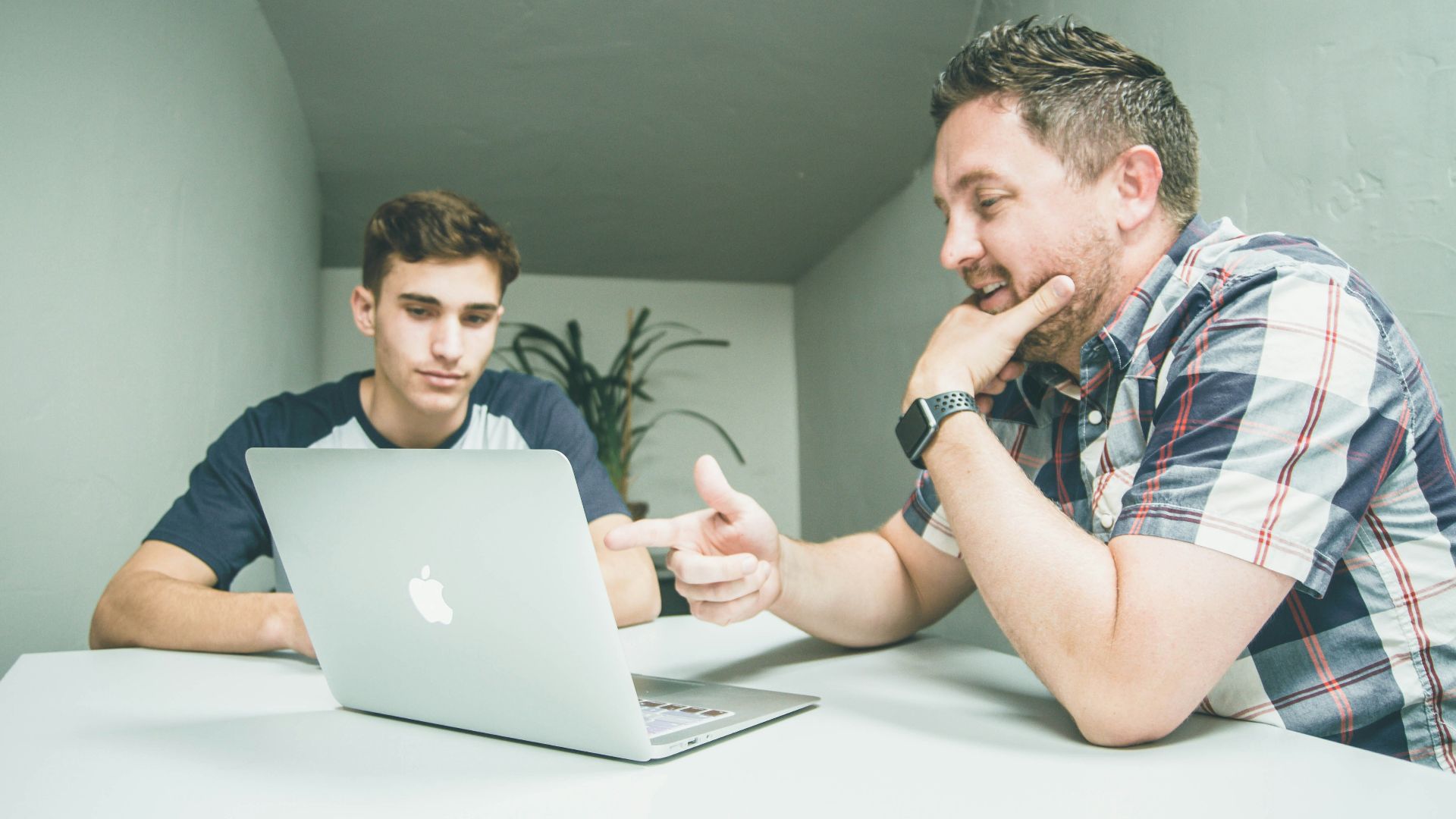 man wearing white and black plaid button-up sports shirt pointing the silver MacBook