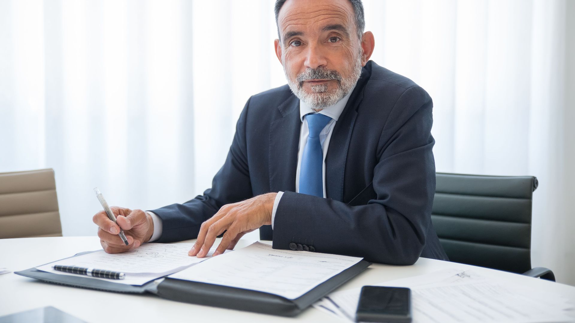 Elderly businessman with a beard signing documents in a modern office setting.