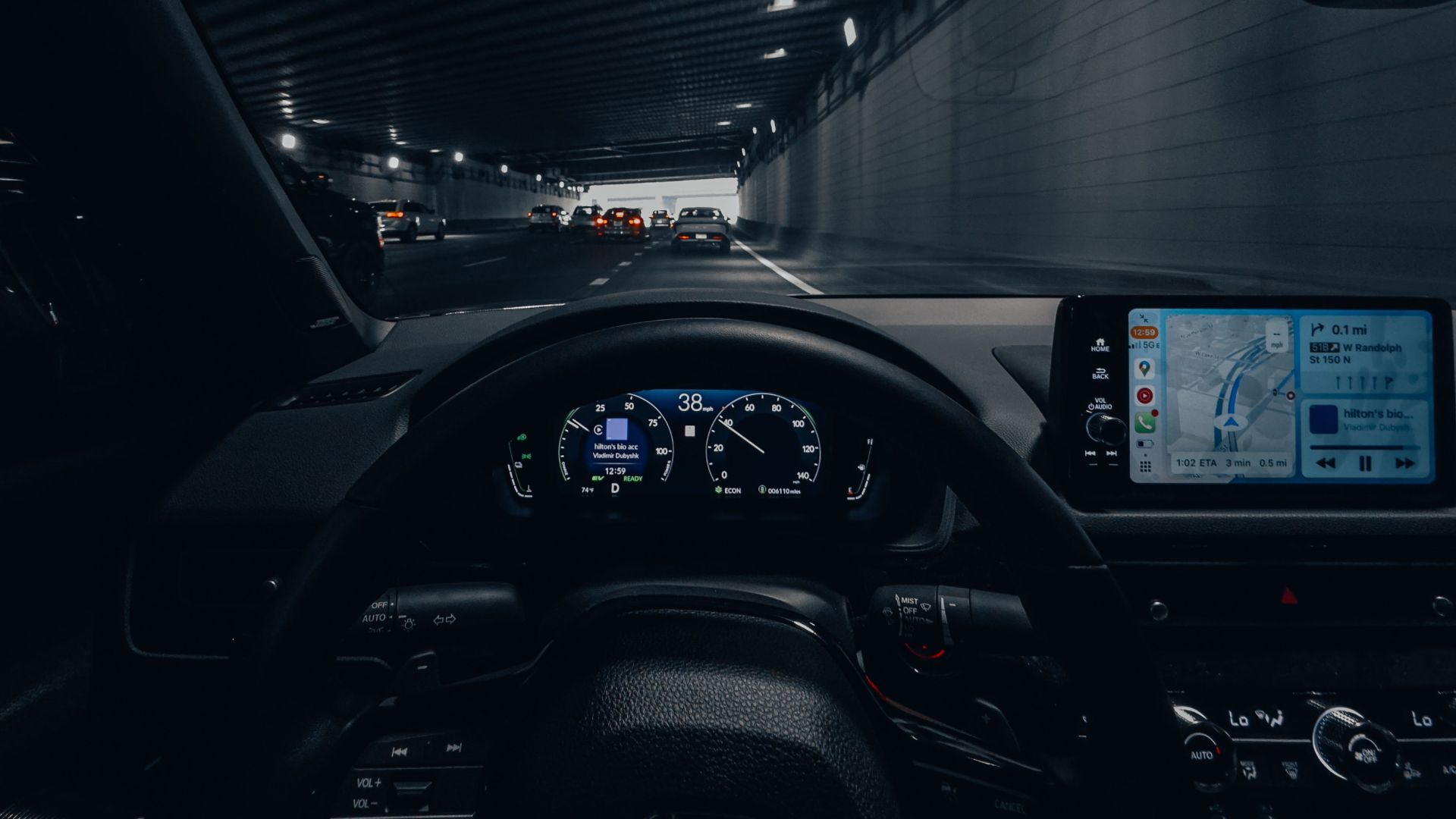 View from car dashboard driving through an illuminated city tunnel at night.