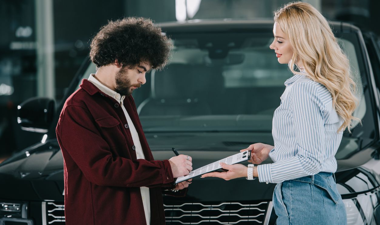 blonde car dealer holding clipboard while curly man signing contract  