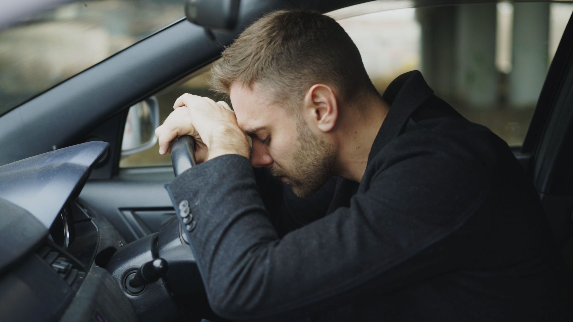 A man showing stress while sitting in a car, head on steering wheel.