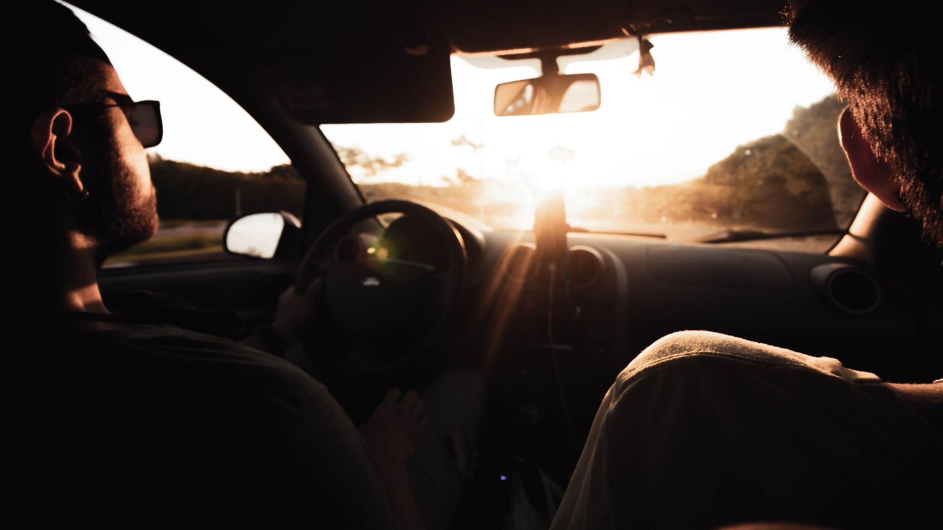 Two men driving in a car interior, enjoying a scenic view with sunlight streaming through the windshield.