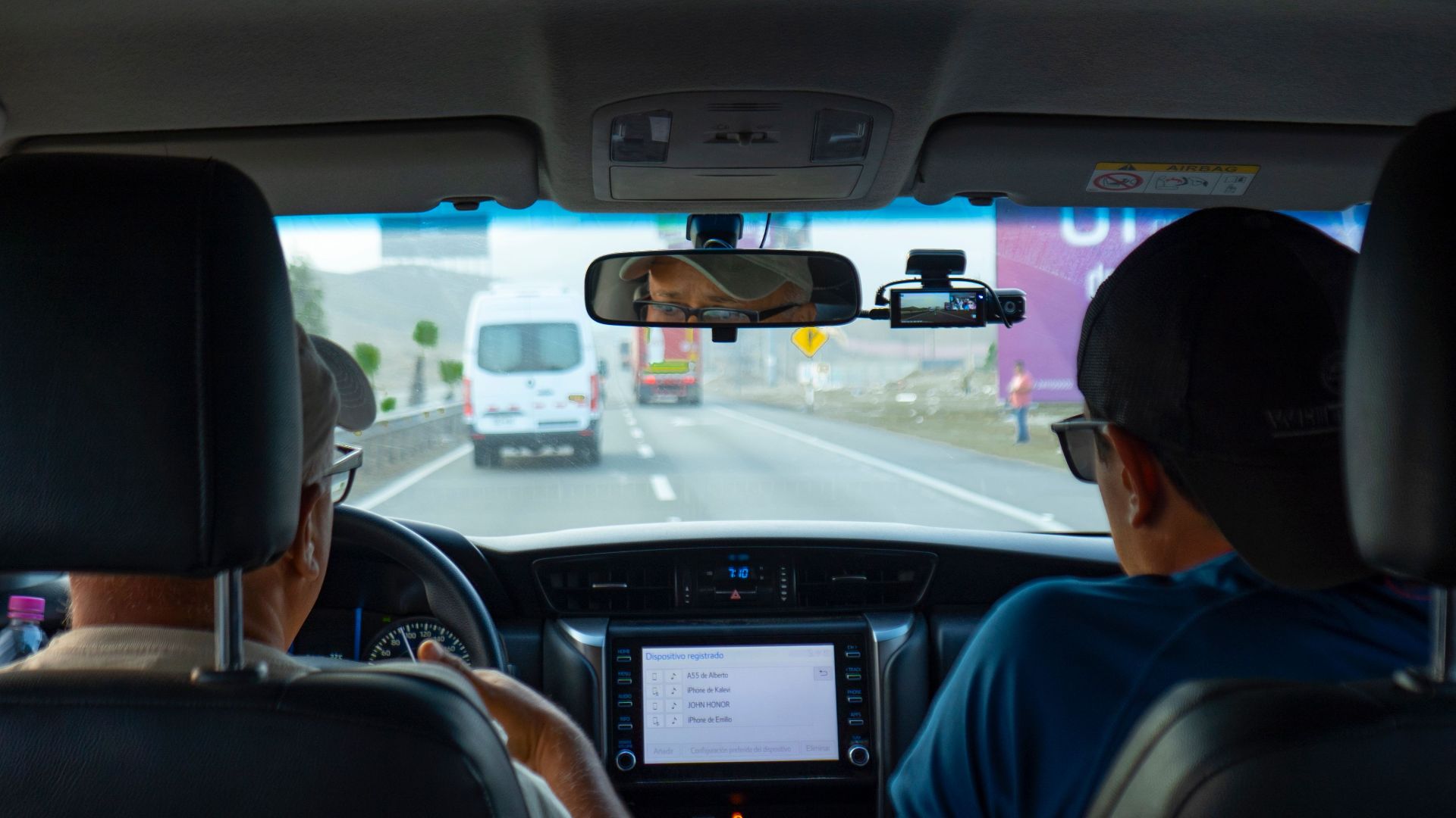 Interior of a car with two people driving on a highway, focusing on dashboard and rear view mirror.
