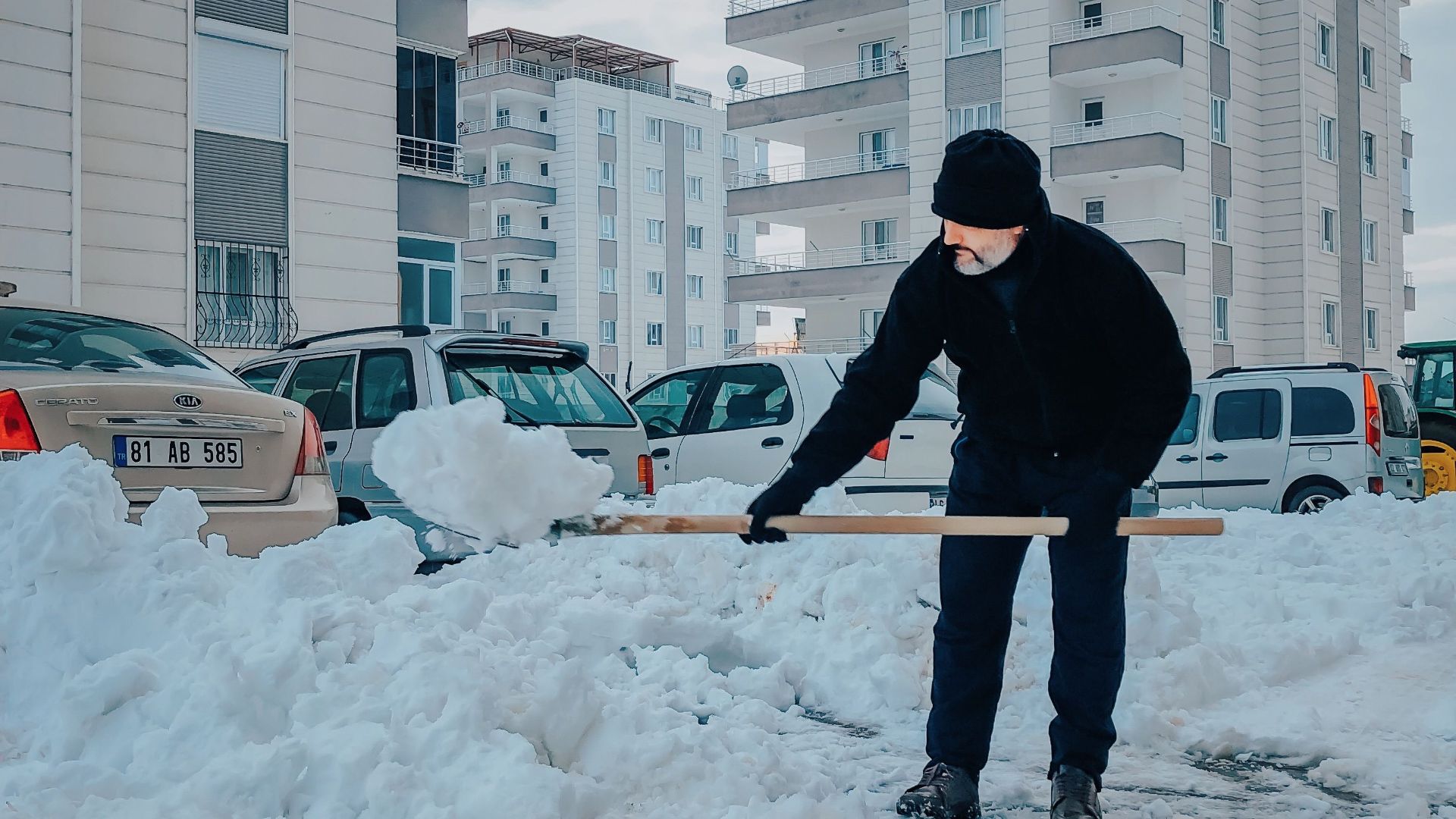 Adult shoveling snow near cars and apartment buildings in winter.