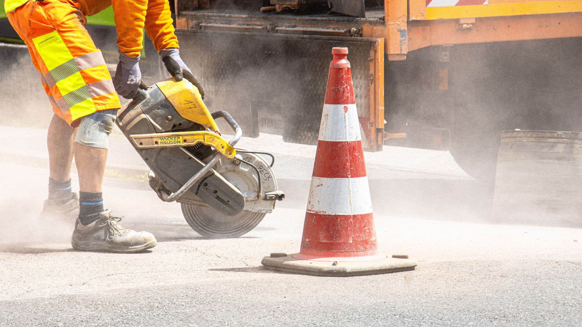 A construction worker cutting asphalt with a large saw next to a traffic cone on a street.