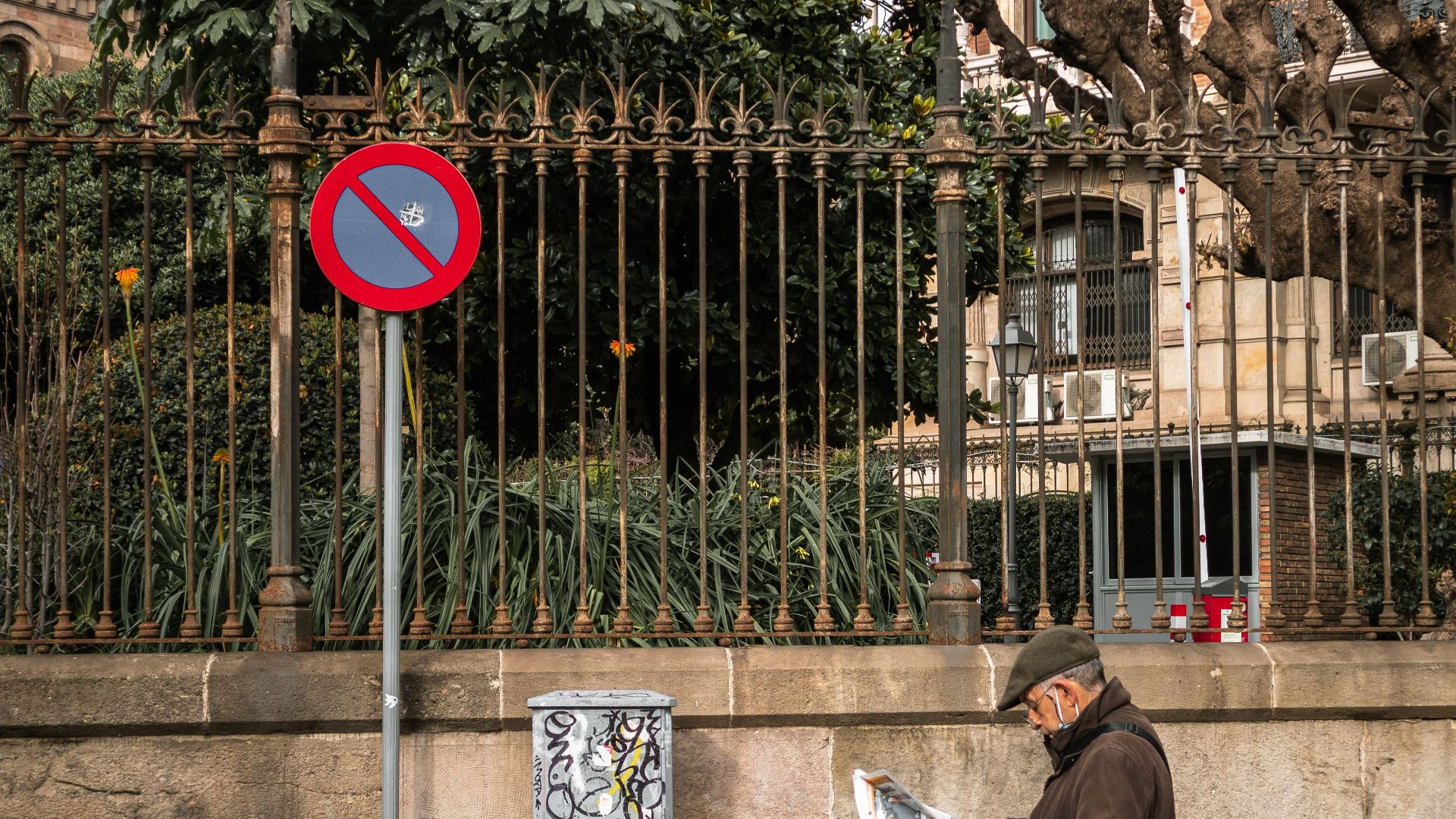 An elderly man reads a newspaper while standing on a Barcelona street, next to a 'no parking' sign.