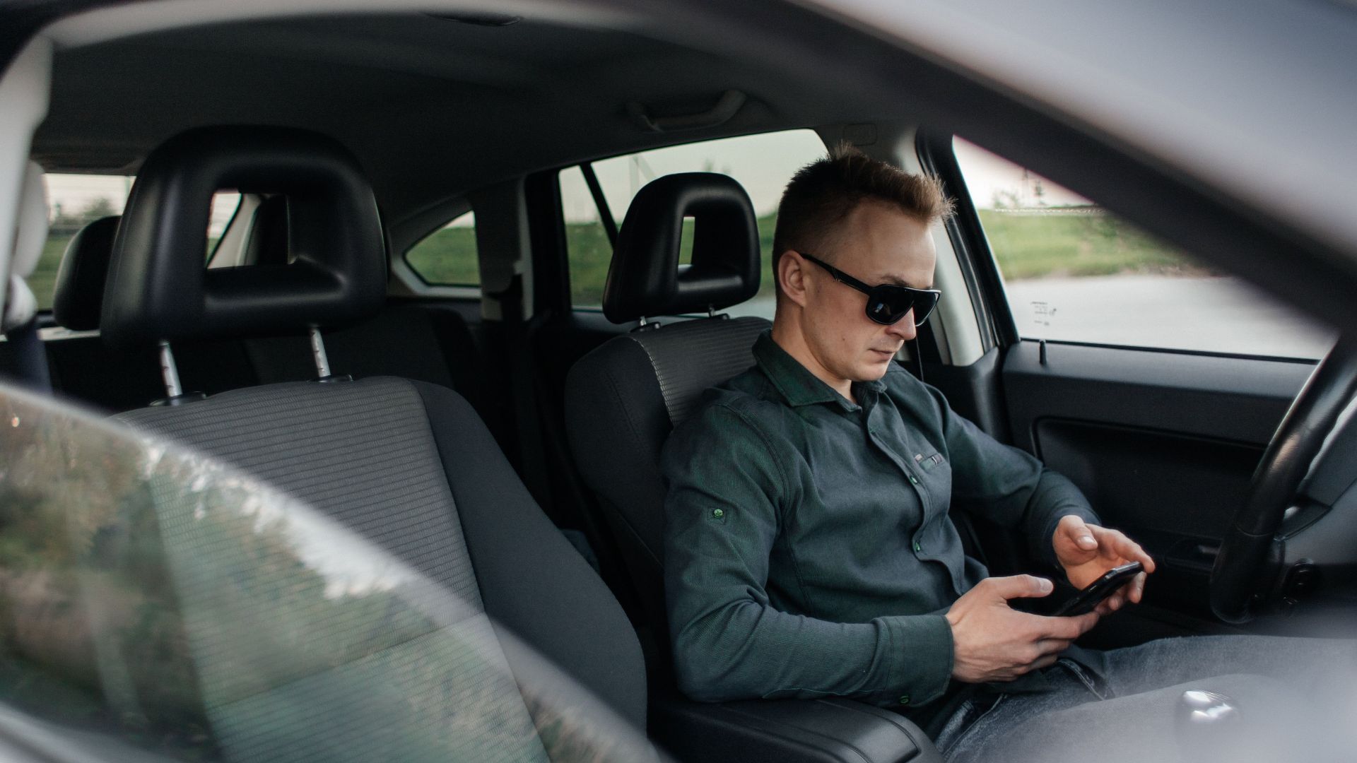 Young man using smartphone inside a parked car, wearing sunglasses.