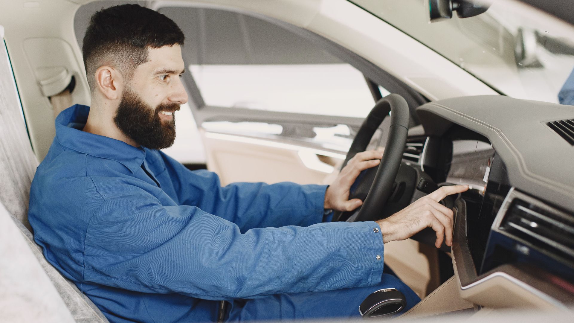 Mechanic in blue coveralls interacts with car dashboard, smiling and focused.