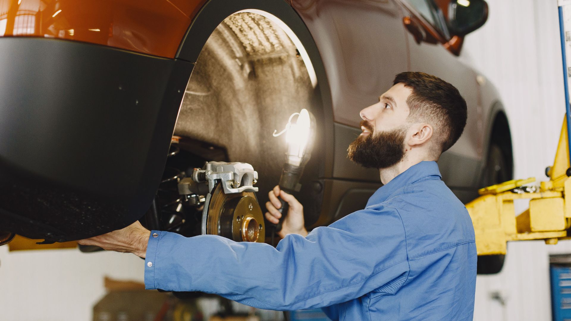 Professional mechanic in blue coveralls inspecting car brakes in a well-lit garage.