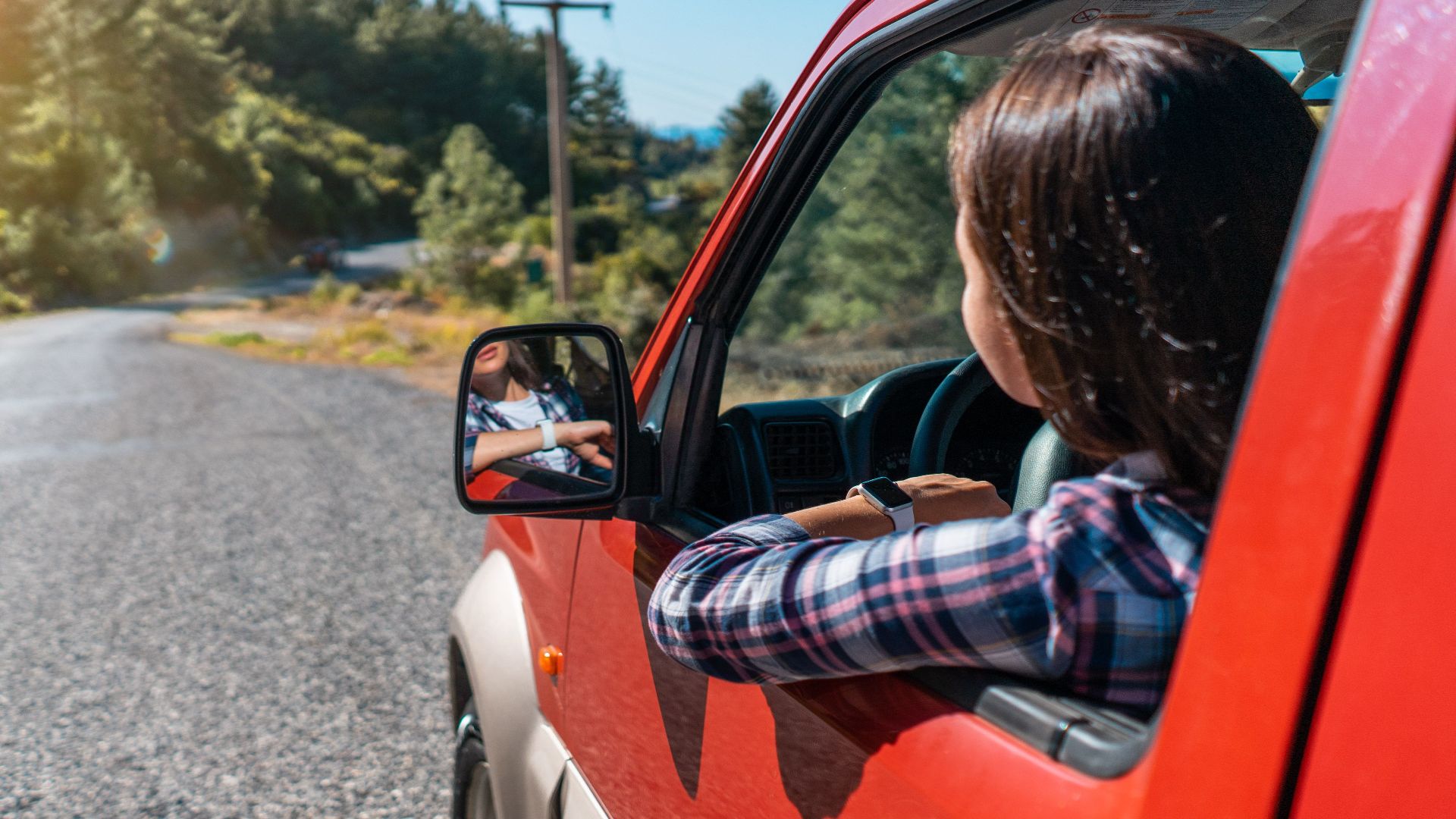 Back view of unrecognizable female driver looking out of window while riding red automobile on asphalt road near green trees