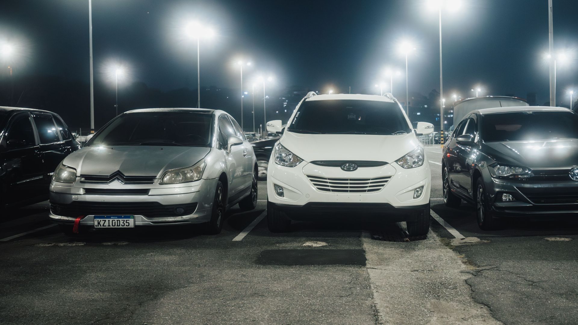 Night scene in a well-lit parking lot featuring various parked cars and street lamps.