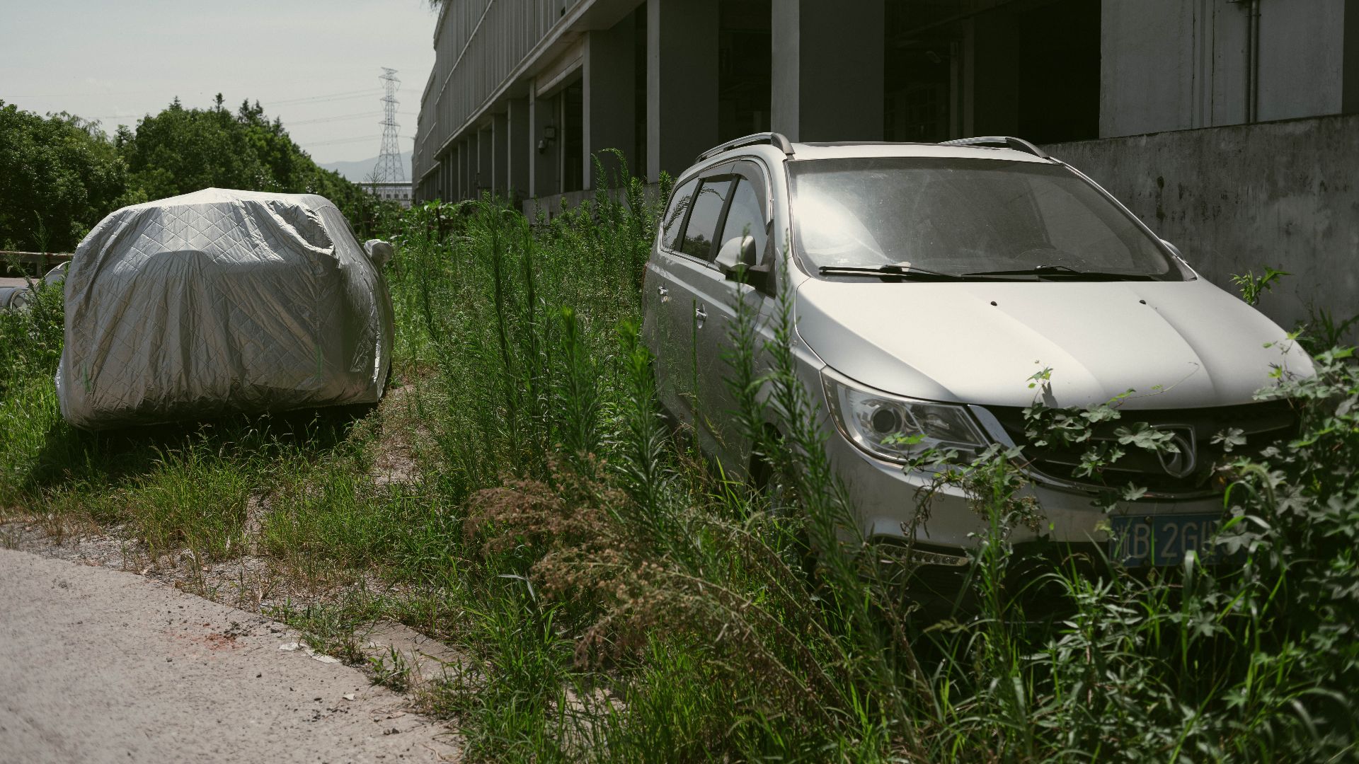Two abandoned SUVs covered in dust and foliage near an industrial building, signifying neglect.