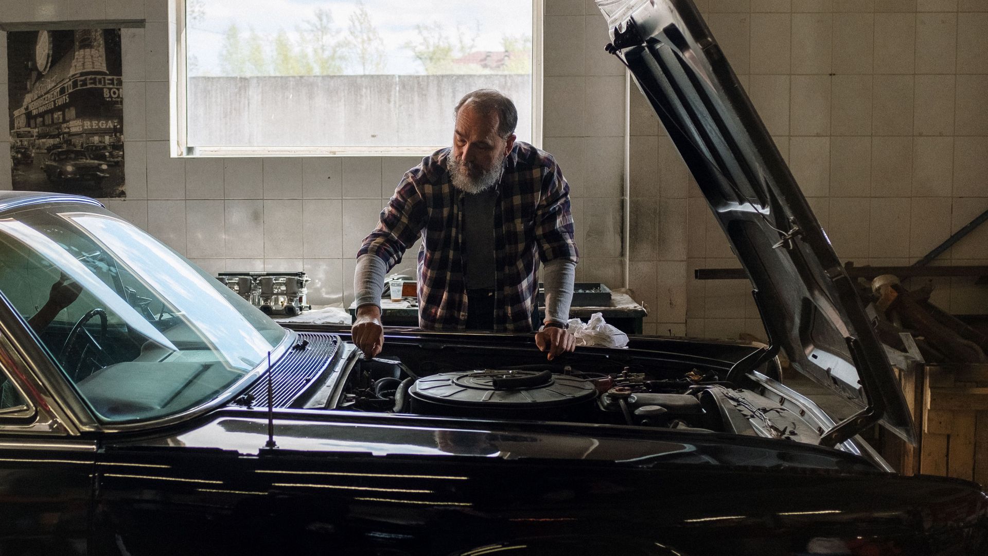 A bearded mechanic examines a classic car engine in a well-lit auto repair shop.