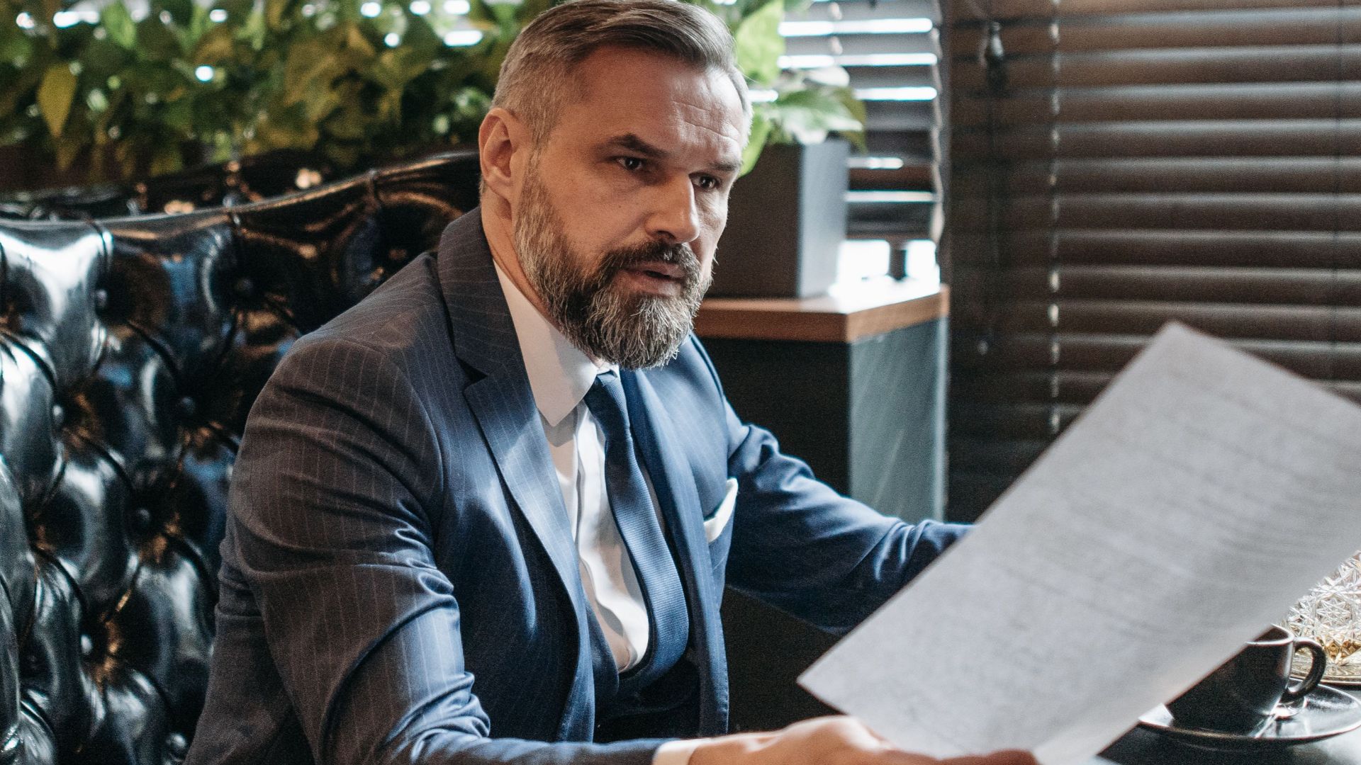 Elderly businessman in a suit analyzing paperwork in a modern office with plants.