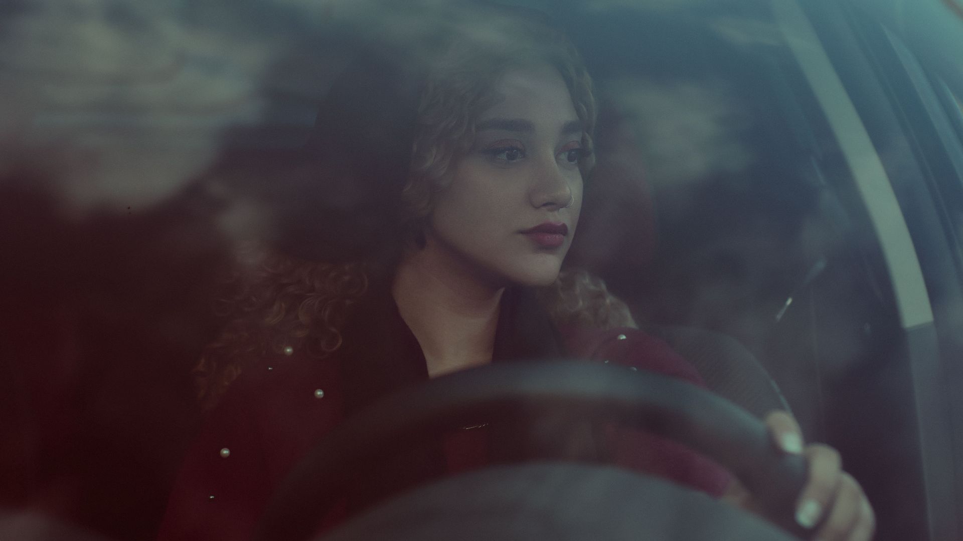 A young woman with curly hair focuses on driving through the city, reflecting her thoughtful expression.