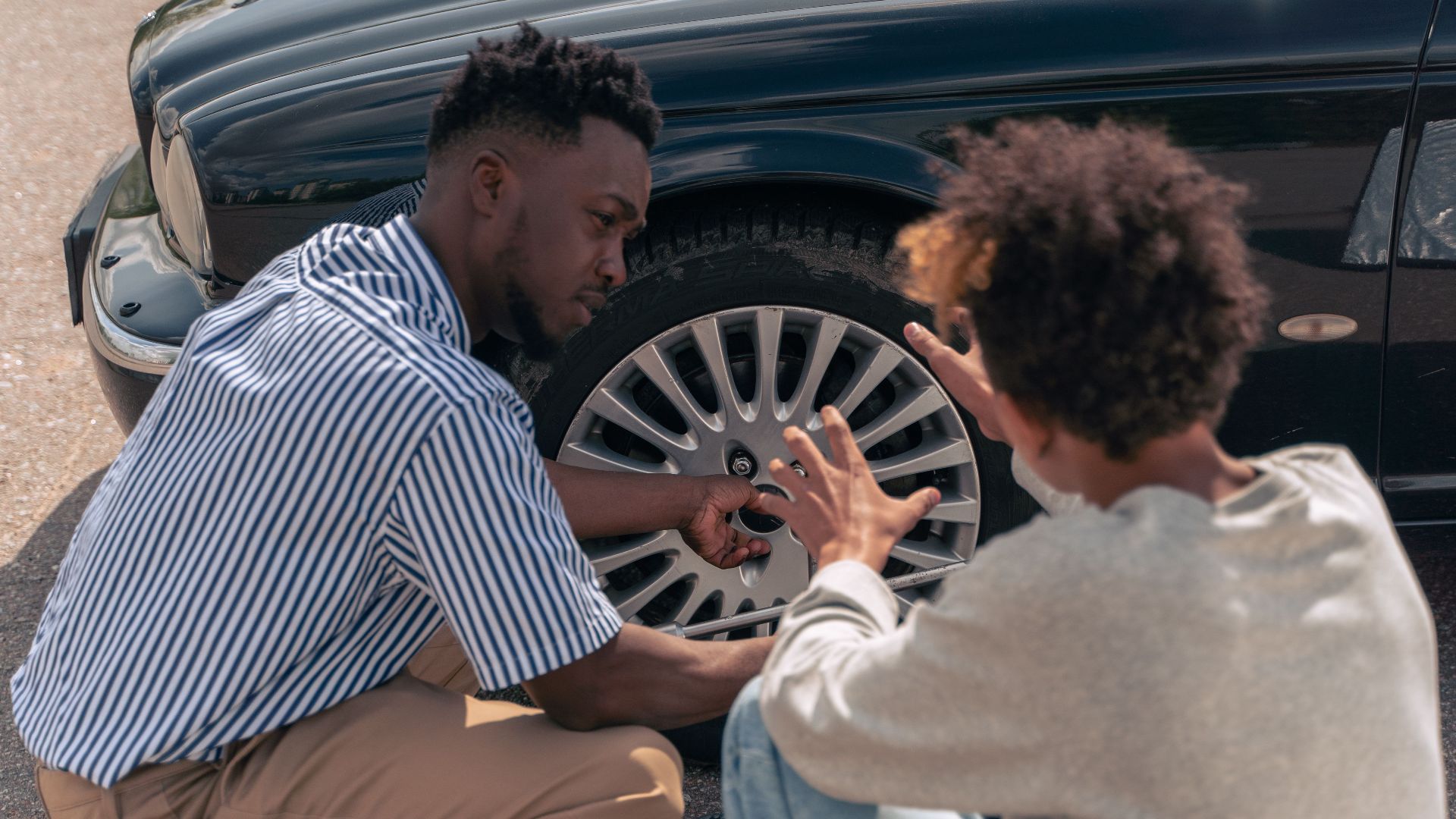 A father instructs his teenage son on how to change a car tire outdoors.