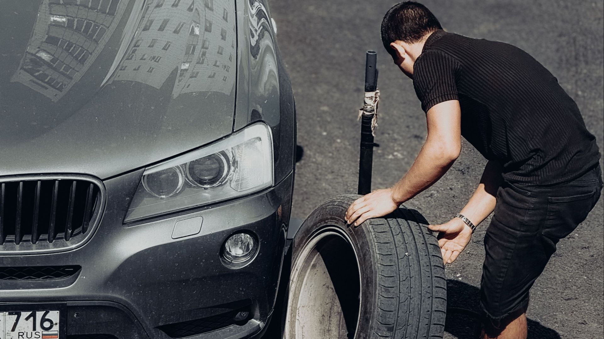 A young man changes a car tire on an outdoor road under the bright sunlight, highlighting the DIY task.
