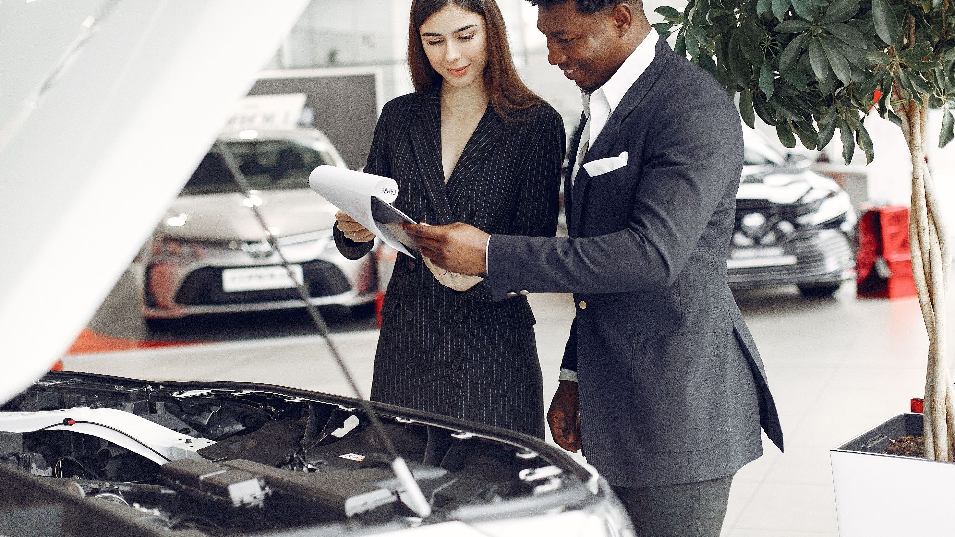 Confident smiling successful black businessman in classy formal suit standing near open motor hood in car showroom and discussing car characteristics with gorgeous female dealer