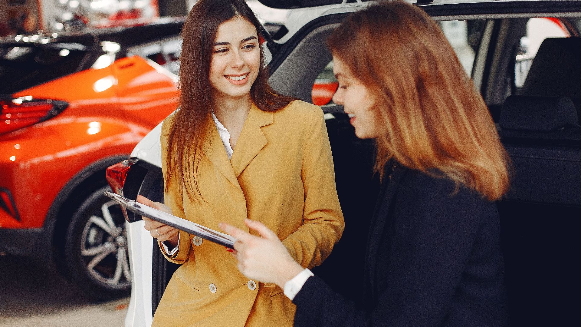 Positive smiling agent in trendy formal light brown suit discussing with happy excited female customer details of contract while leaning on car in showroom