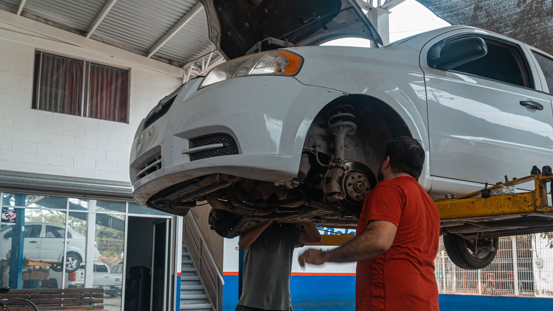 Two mechanics inspecting a white car on a lift inside a garage workshop.