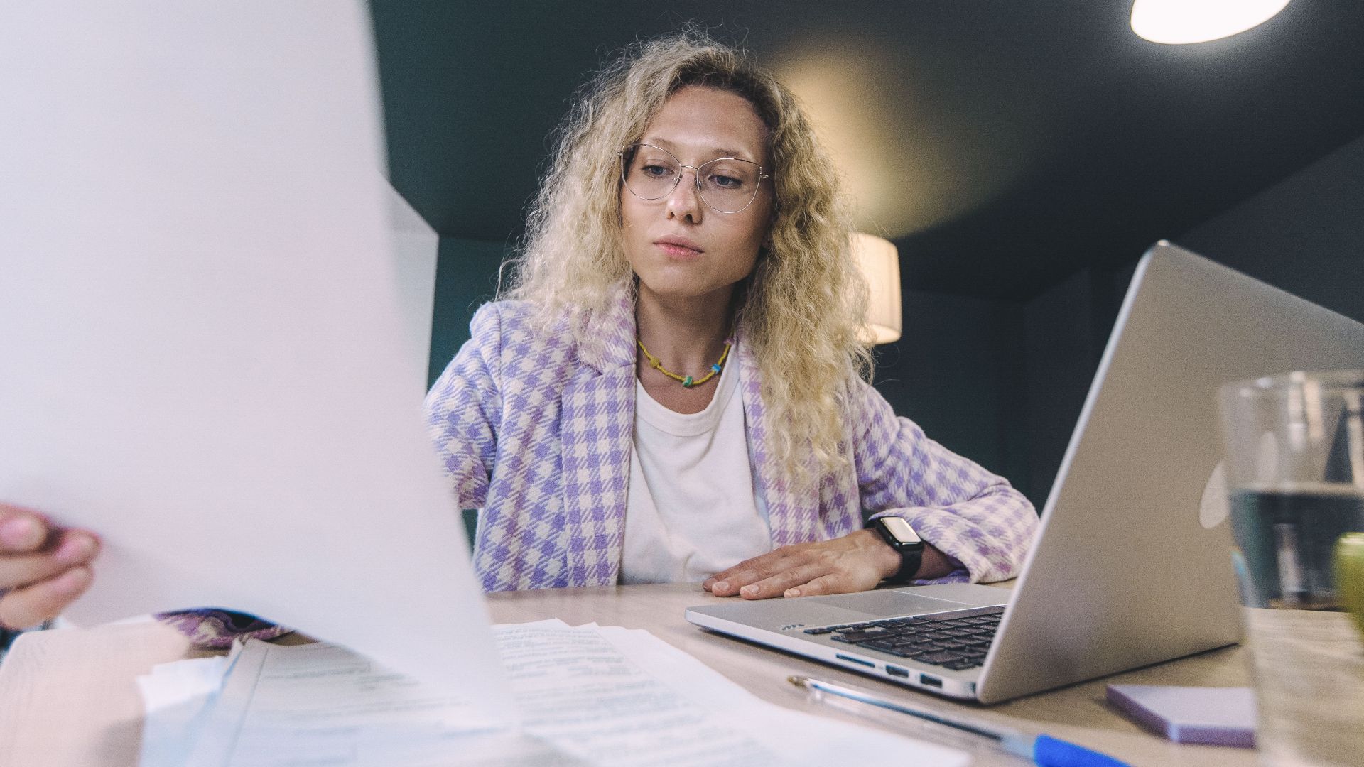 Woman in an office environment reviewing documents with focus, surrounded by technology.