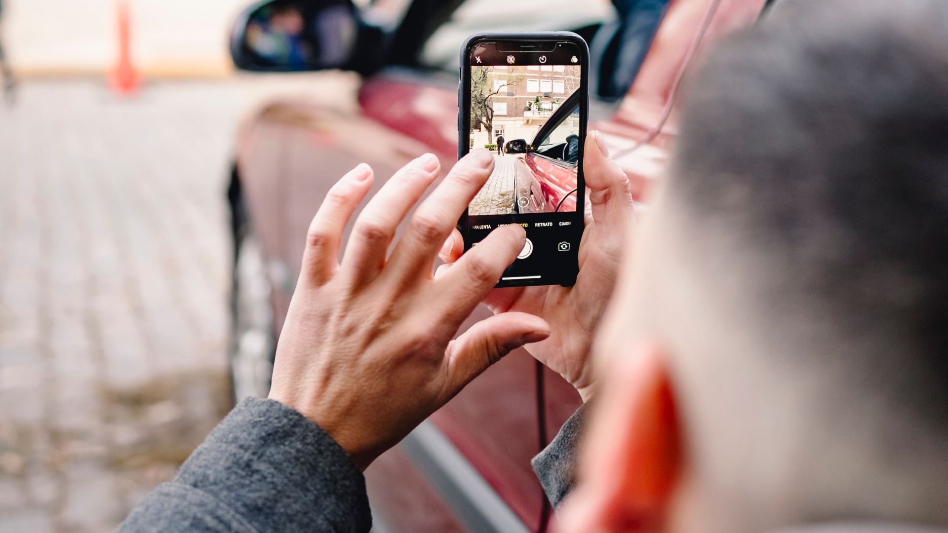 Man using smartphone to take pictures of an urban scene beside a red car.