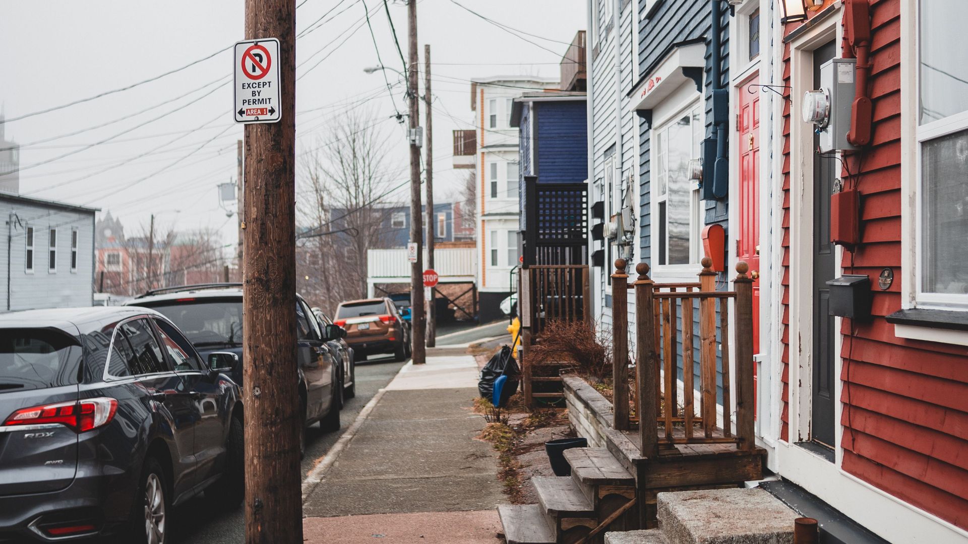 Straight sidewalk between colorful residential house with stone steps and roadside with cars on street with power lines in town