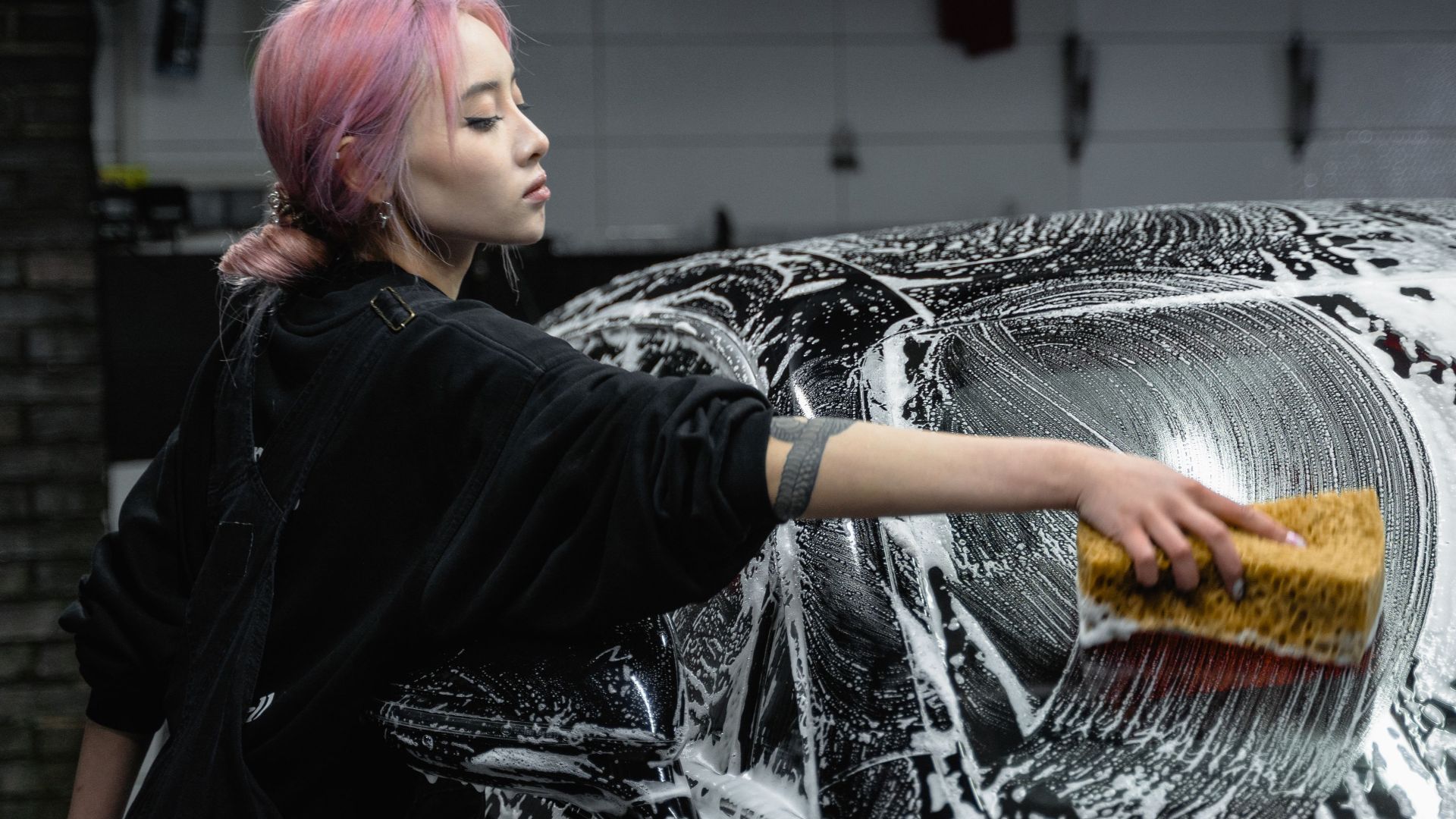 A young woman cleans a black car with a sponge and soap in a garage setting, showing care and attention.