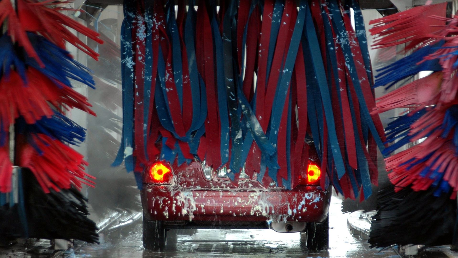 Red car being cleaned in an automated car wash with colorful brushes and soap suds.