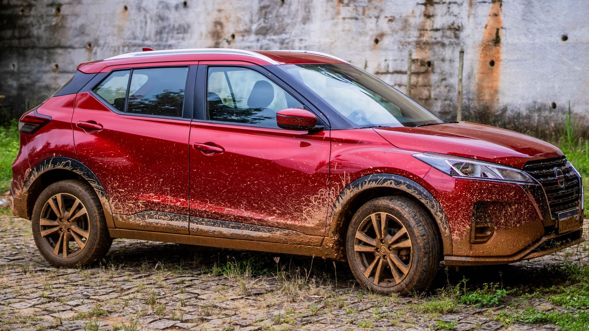 A red Nissan SUV with mud splatters seen parked on cobblestone.