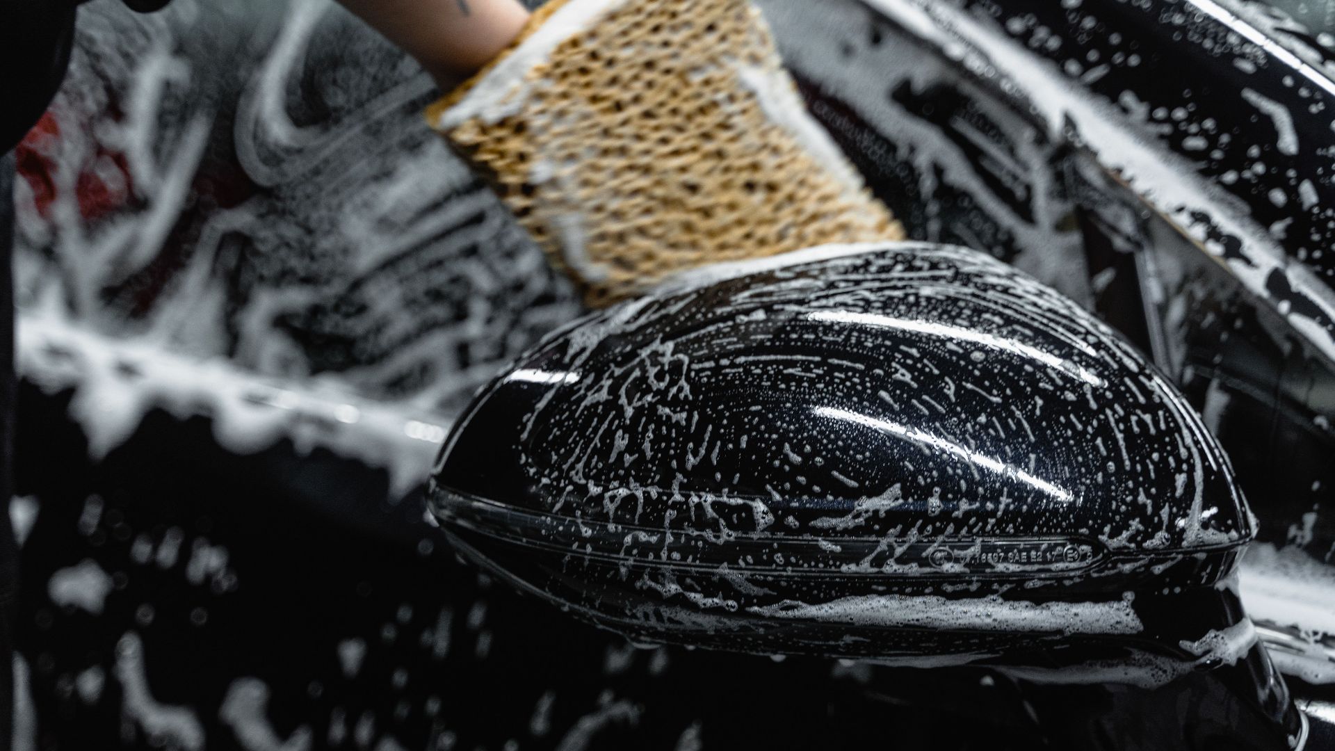 Hand scrubbing a car mirror with a sponge covered in soap at a car wash.