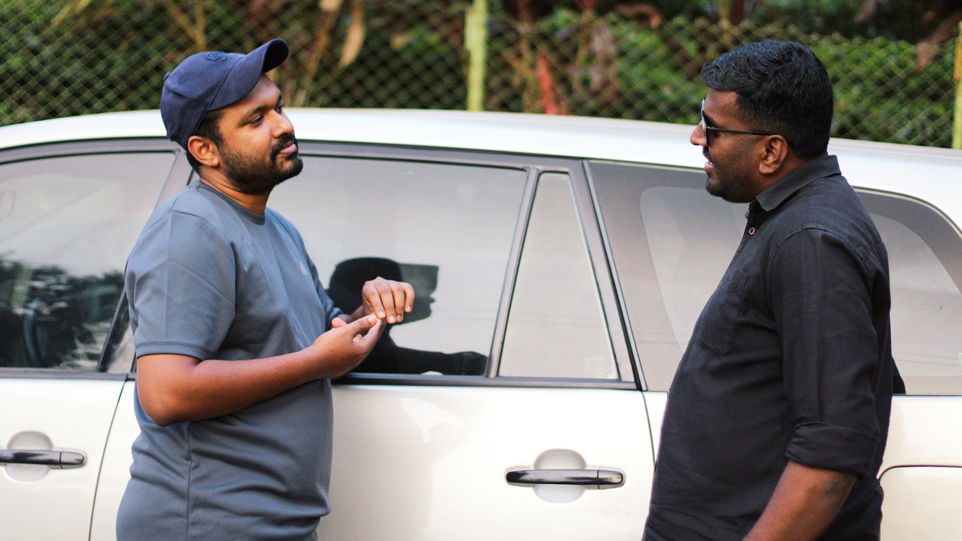 Two men having a casual discussion beside a car in an outdoor setting.