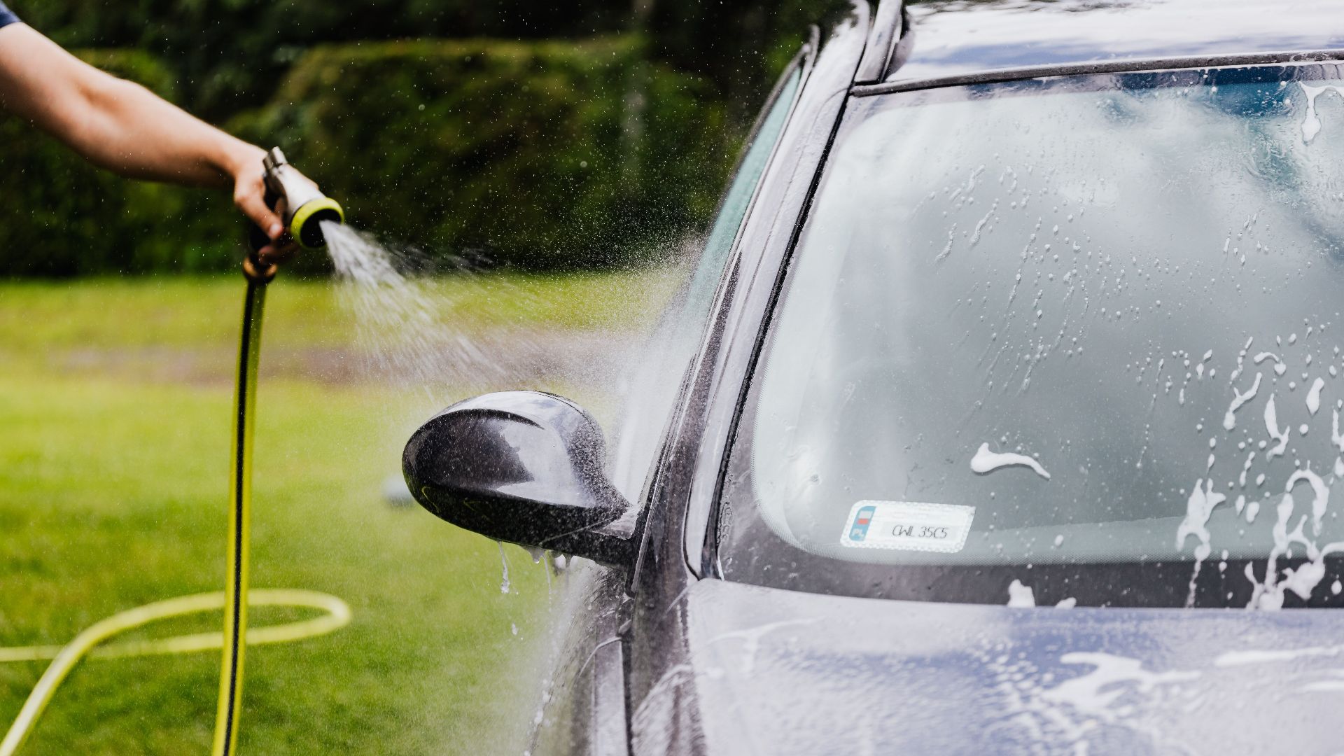 Person washing a car with a hose outdoors, creating suds on a sunny day.