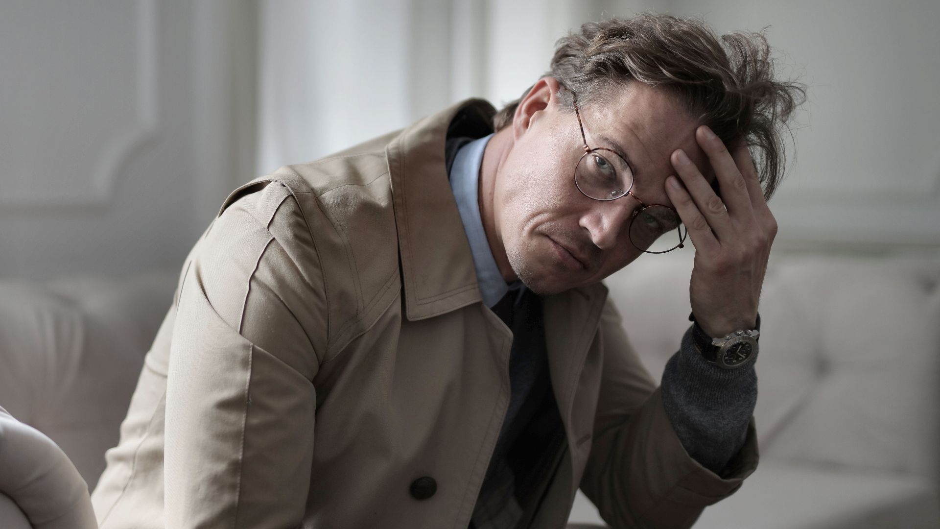 Pensive adult brown haired man in eyeglasses and trench sitting on couch and touching head while looking at camera