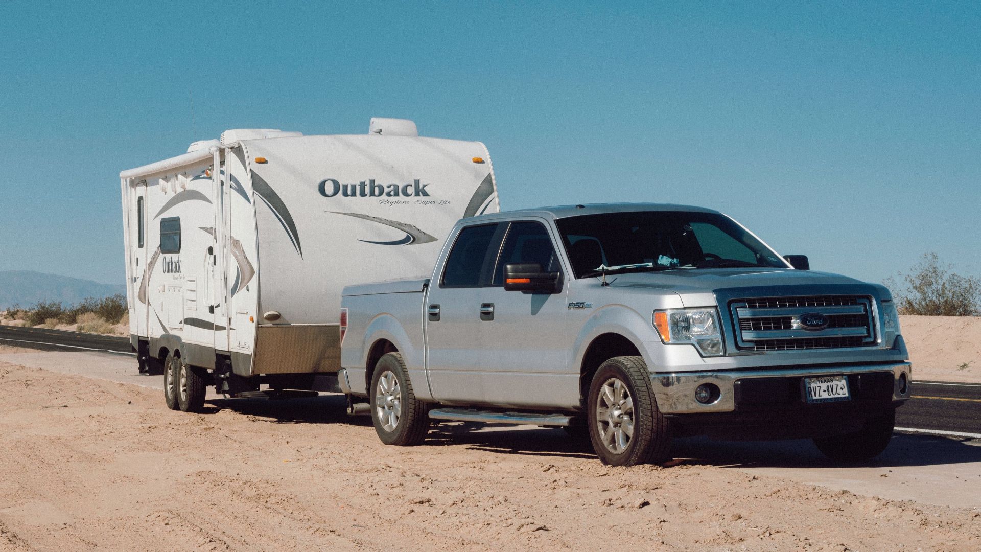 Silver truck towing an Outback trailer on a desert road under clear blue skies.