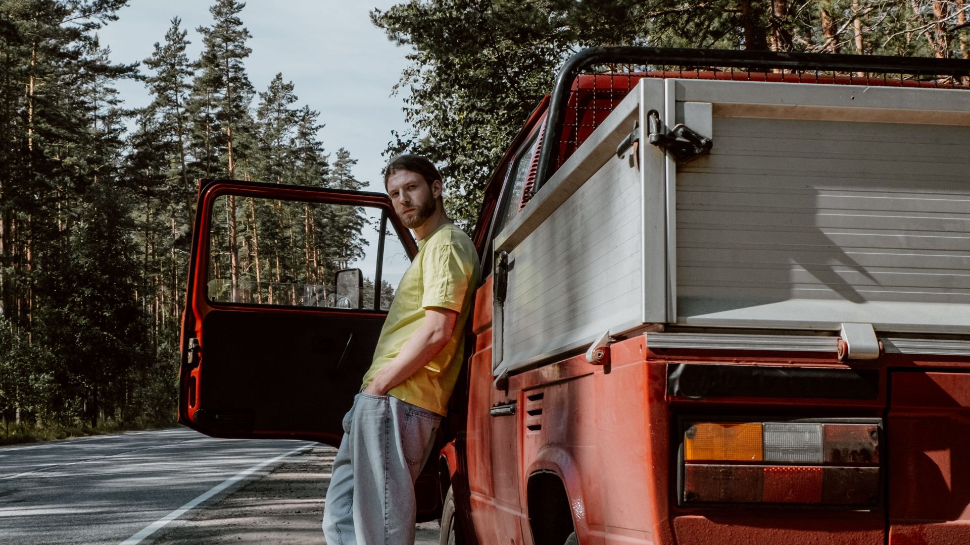 A man stands by his red pickup truck on a quiet country road surrounded by tall trees.