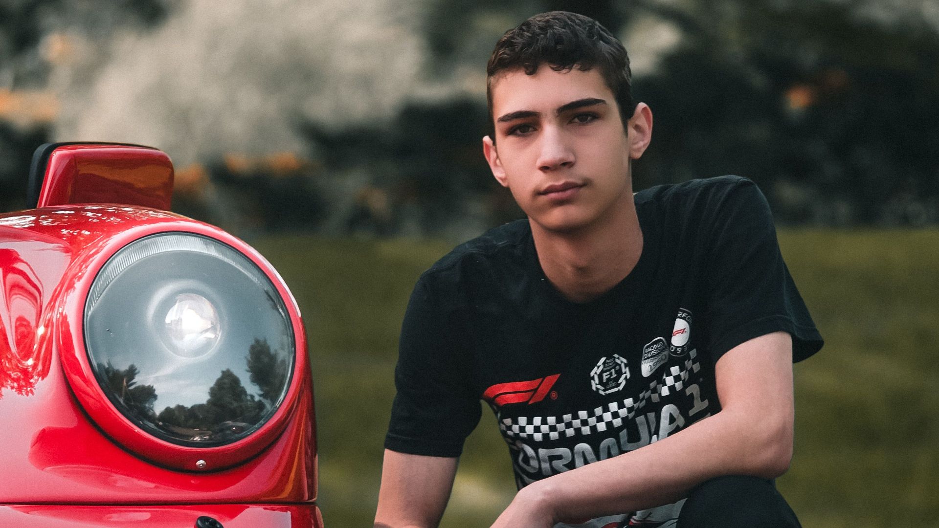A teenager in casual wear kneeling by a vintage red sports car in a park setting.