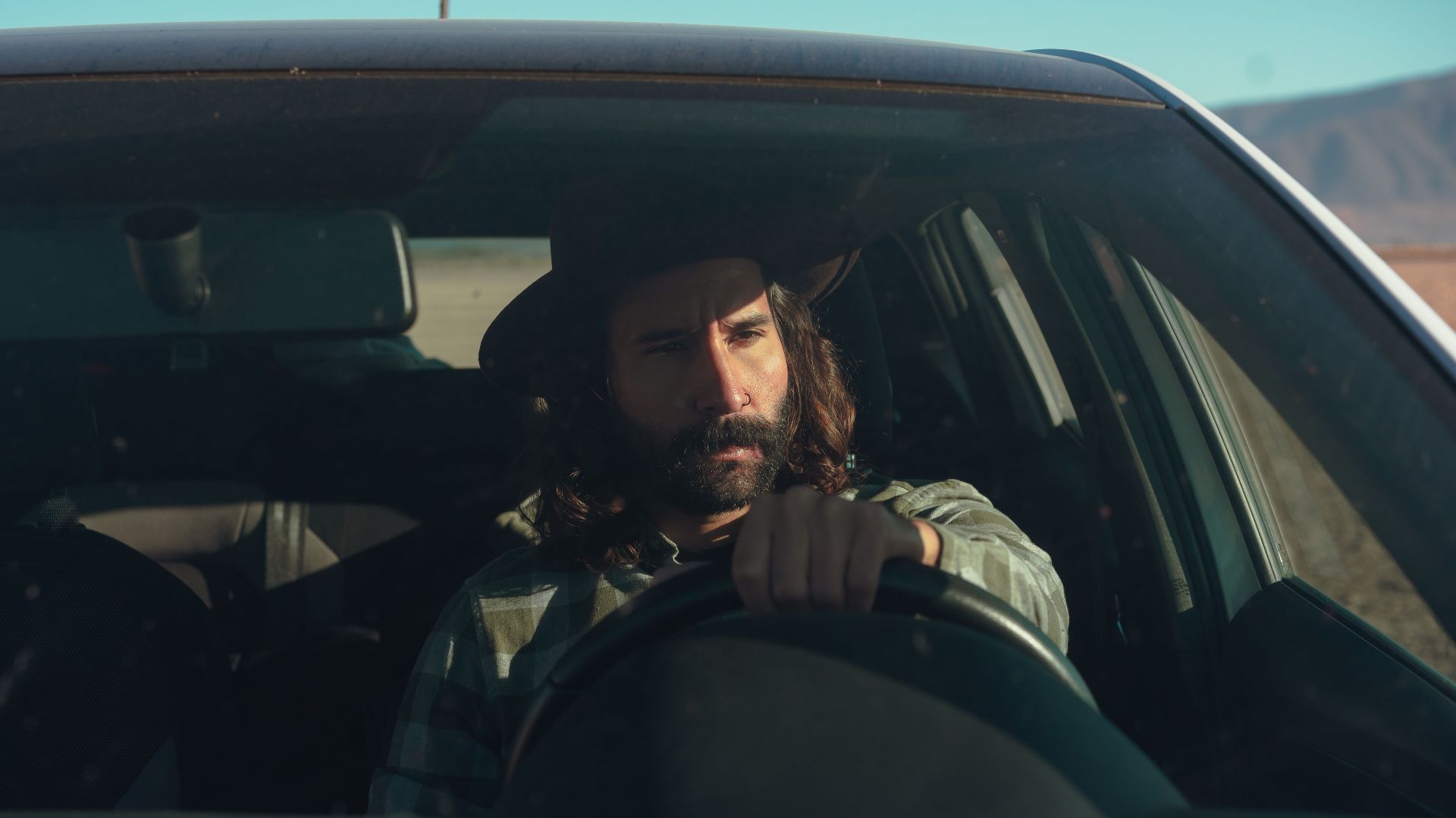 A bearded man in a hat driving a car through a sunny desert landscape, conveying a sense of adventure.
