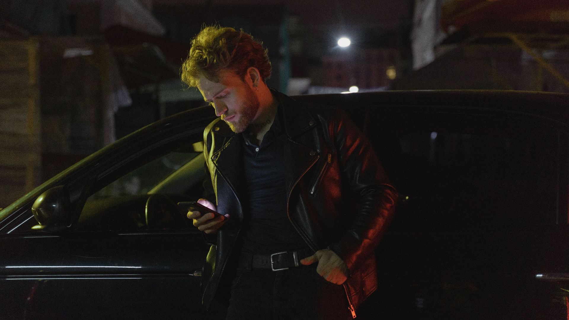 Caucasian man stands by a car at night, using his phone. Retro 80s style with a moody atmosphere.