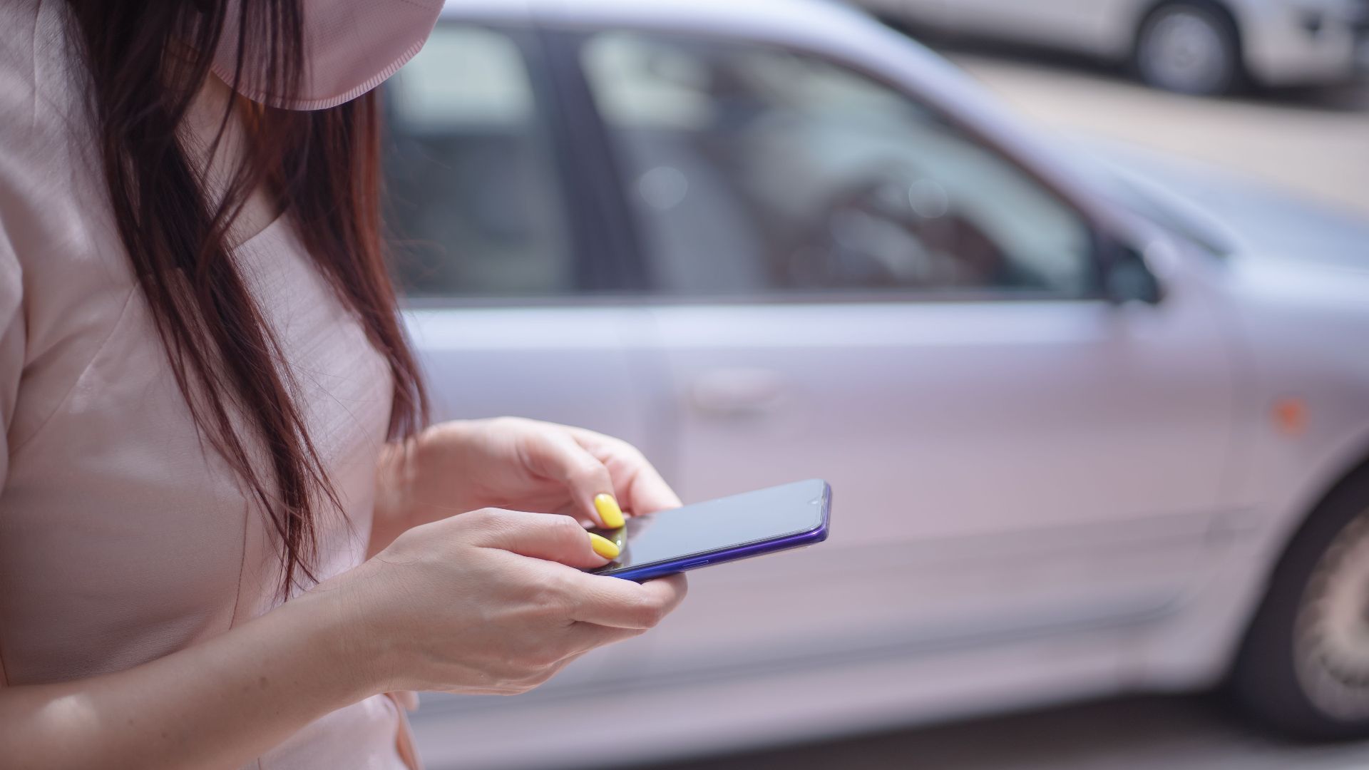 A close-up of a woman texting on a smartphone near a parked car outdoors.