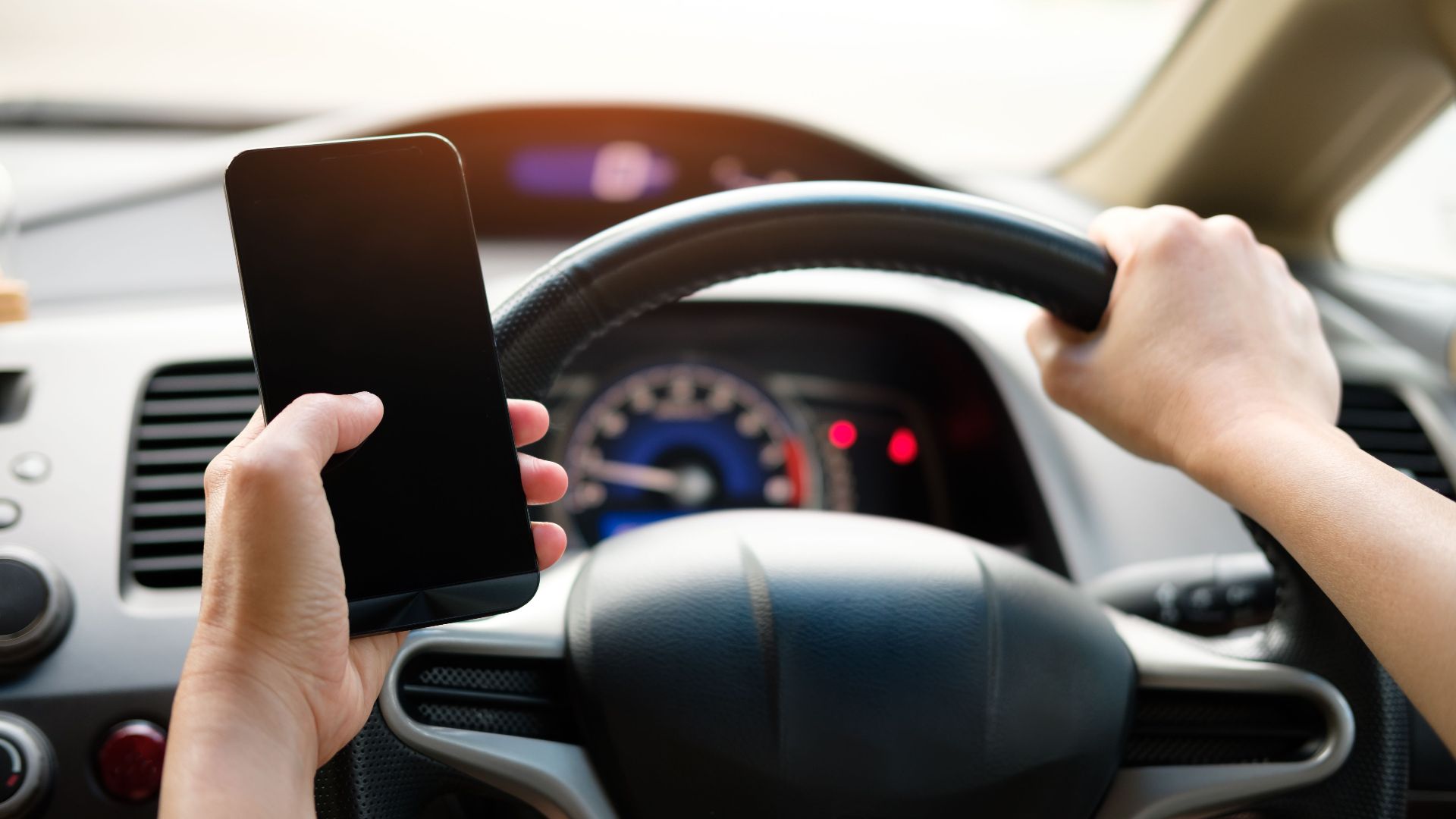 A driver uses a smartphone while steering a vehicle, showcasing distracted driving risks.