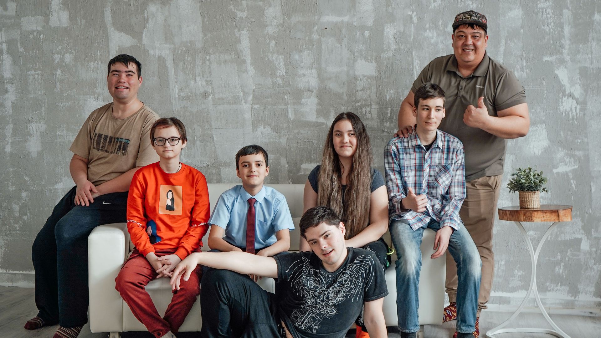 Group of seven people posing casually in a stylish indoor setting with a concrete wall background.