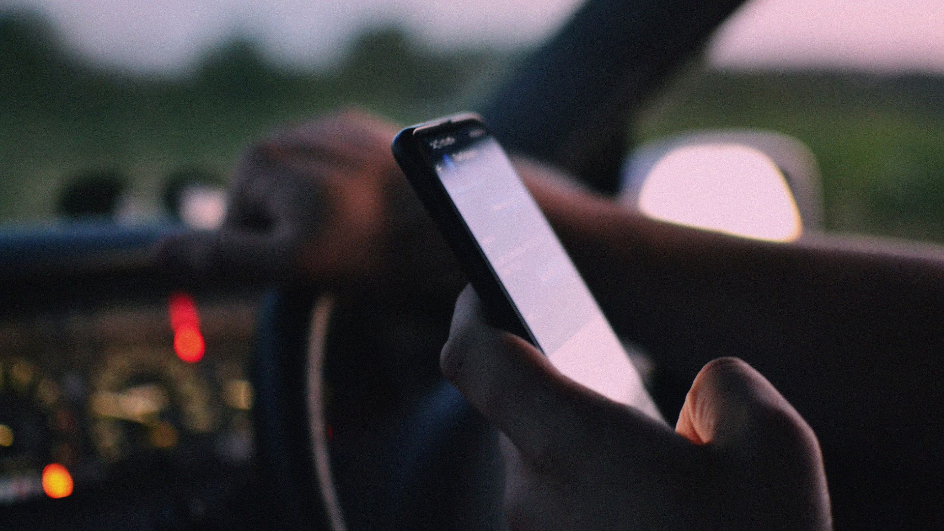 Close-up of a person using a smartphone while driving in a car at dusk.