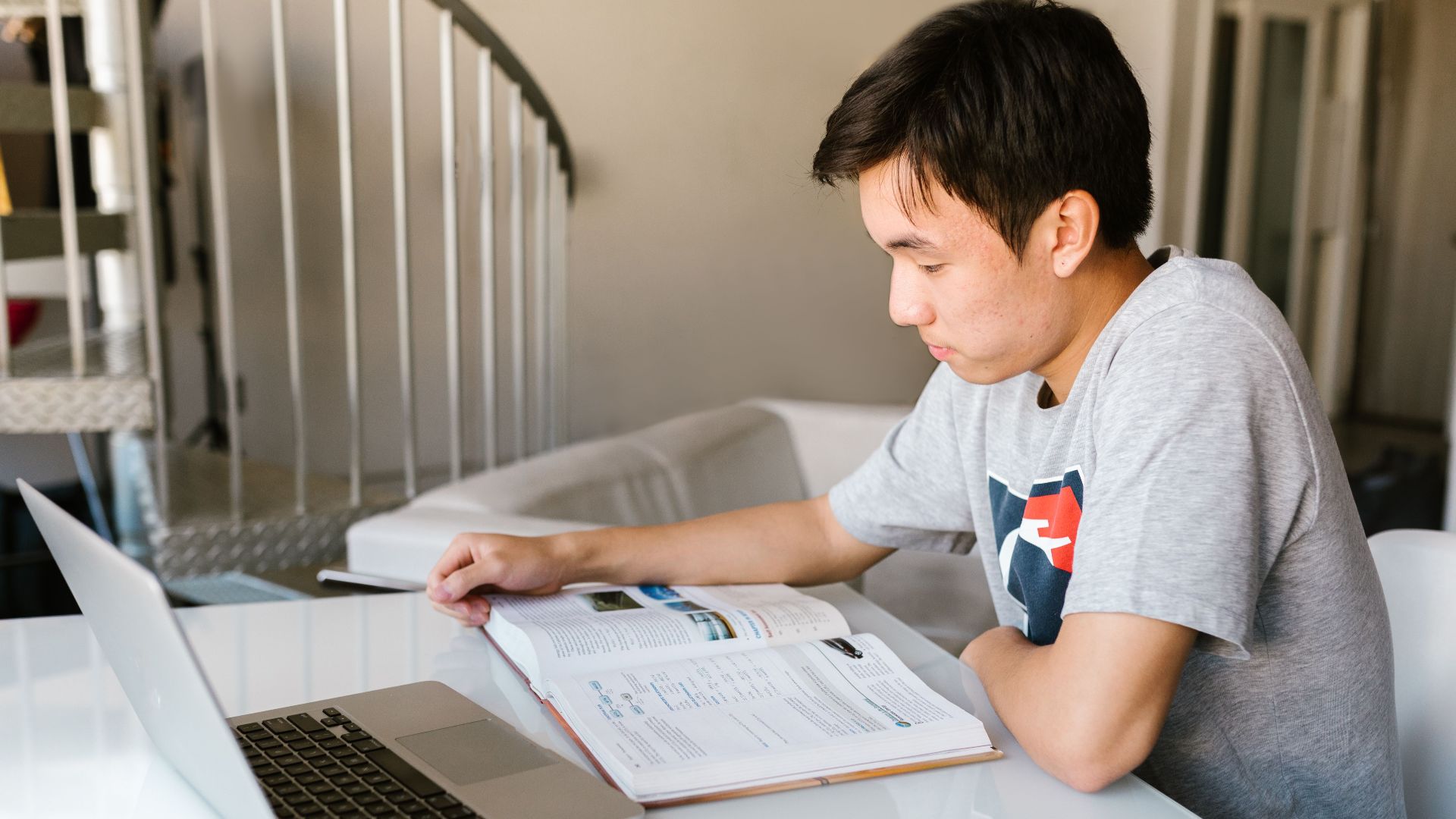 Teenage boy focuses on homework using a laptop and textbook indoors.