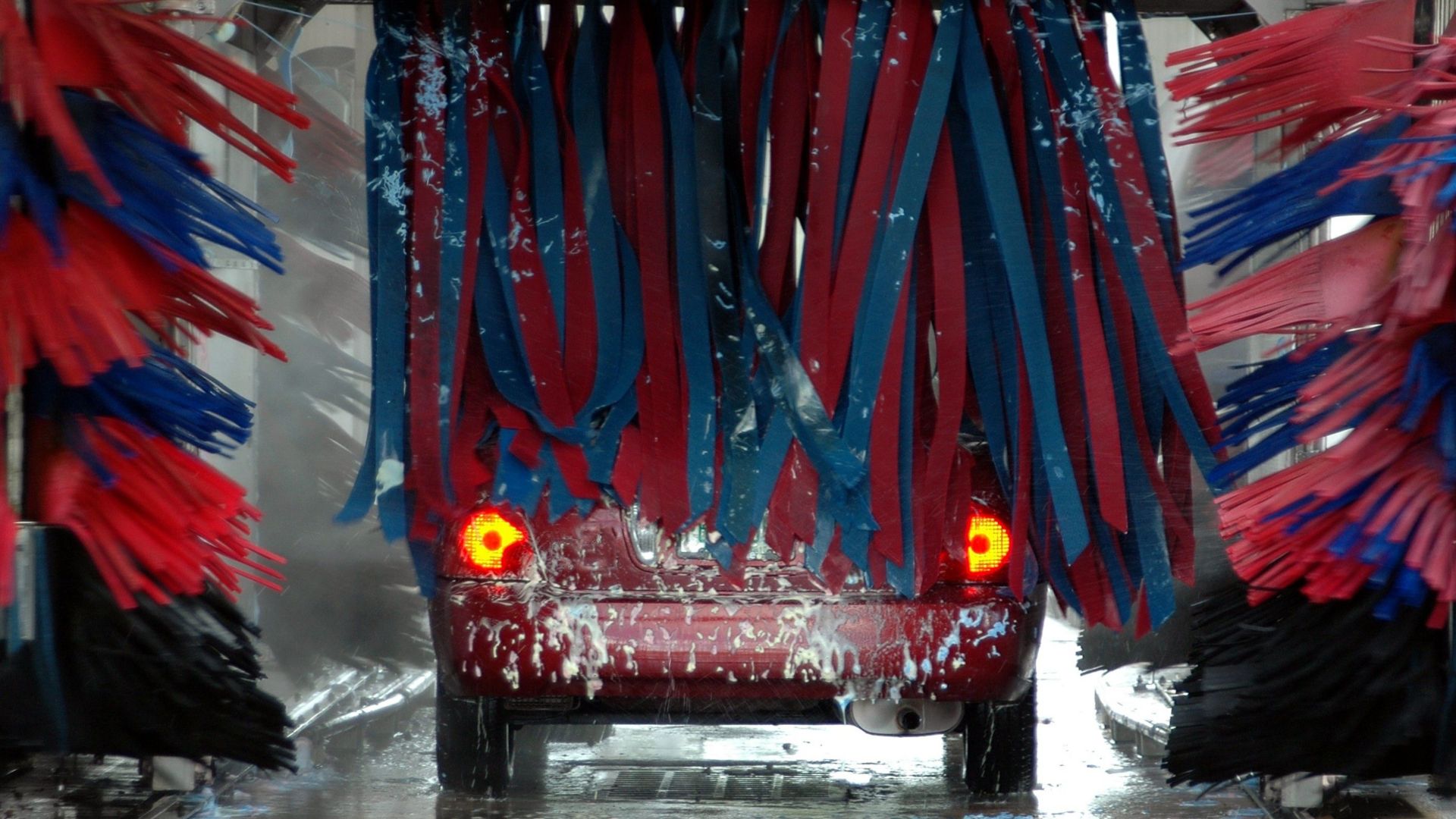 Red car being cleaned in an automated car wash with colorful brushes and soap suds.